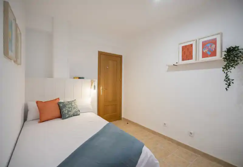 Chambre lumineuse et bien décorée avec un lit double adossé à une tête de lit blanche, coussins colorés et un plaid bleu-vert. Sol carrelé chaleureux, cadres muraux sobres et plante suspendue pour une atmosphère cosy.