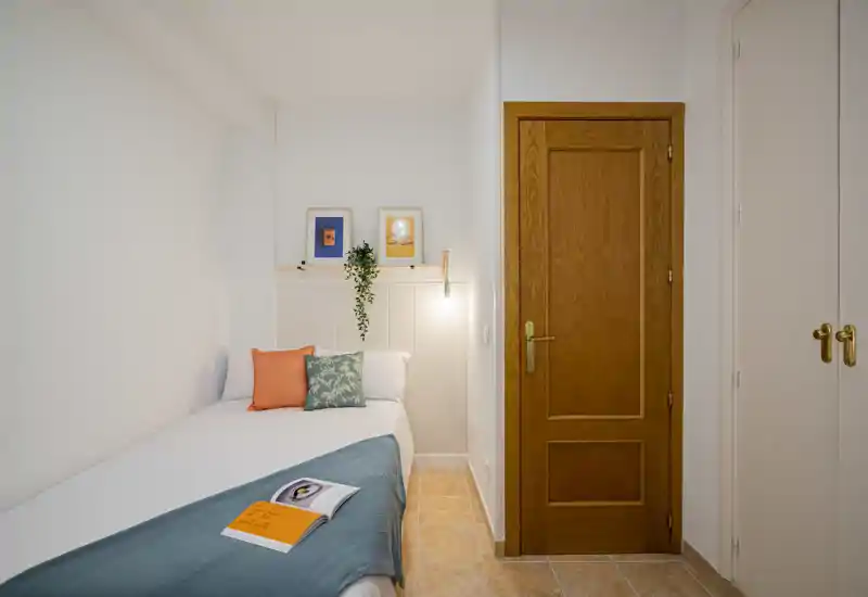 Compact, well-styled single bedroom with a neatly made bed, decorative cushions, framed artwork and soft wall lighting — bright and welcoming.