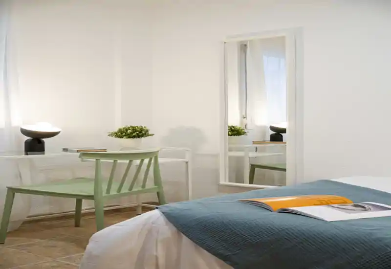 Bright, neatly staged bedroom with a teal bed runner, a green chair and a simple desk by the window; soft natural light and minimal décor create a calm, welcoming atmosphere.