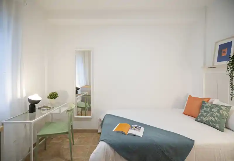 Bright, minimal bedroom with a neatly made double bed, colorful cushions and a bedside desk by the window — ideal for a restful stay or working corner.