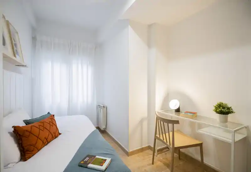 Small, bright bedroom with a comfortable single bed, decorative cushions, a desk and a lamp — ideal for a cozy short-stay stay with natural light from the window.