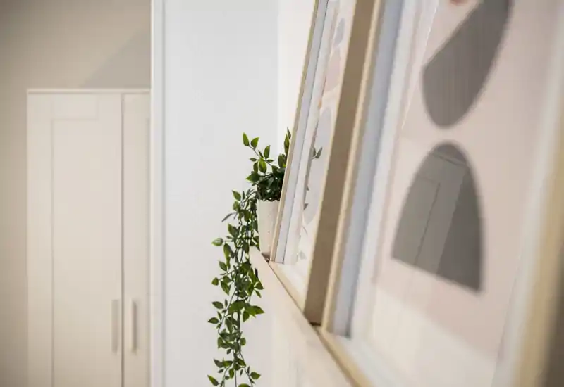 Close-up of decorative framed prints on a narrow shelf with a trailing plant and a blurred cabinet in the background. The image highlights décor details and a calm neutral palette.