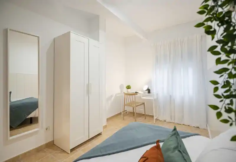 Dormitorio luminoso y ordenado con cama visible en primer plano, armario blanco, espejo de cuerpo entero y un escritorio junto a la ventana con cortina vaporosa; suelo de baldosas y cojines decorativos que aportan calidez.