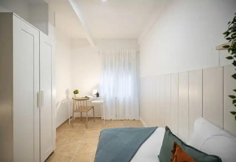 Dormitorio luminoso y bien decorado con cama visible, armario y un pequeño escritorio junto a la ventana. La luz suave y el suelo cálido aportan una sensación acogedora y aireada, perfecto para anuncios de alquiler.
