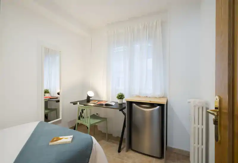 Coin chambre petit et lumineux avec lit bien fait, bureau compact et chaise près de la fenêtre, lampe et mini-frigo — parfait pour un séjour individuel confortable.