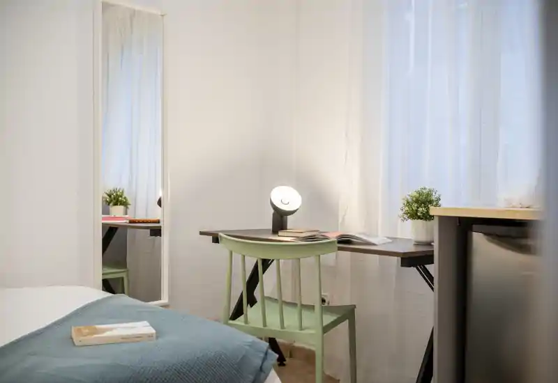 Coin de chambre lumineux et soigné avec un lit bien fait, un petit bureau et une chaise près de la fenêtre, agrémenté d'une lampe et d'une plante.