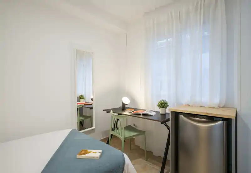 Chambre lumineuse et minimaliste avec un lit bien fait, un petit bureau et un mini-réfrigérateur — parfaite pour une personne ou des courts séjours. La lumière naturelle et le décor sobre offrent une ambiance reposante.