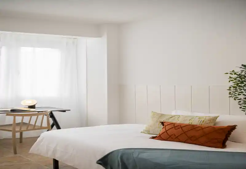 Camera da letto luminosa e minimal con letto ben rifatto, cuscini decorativi e una piccola scrivania vicino alla finestra che creano un'atmosfera moderna e rilassante.