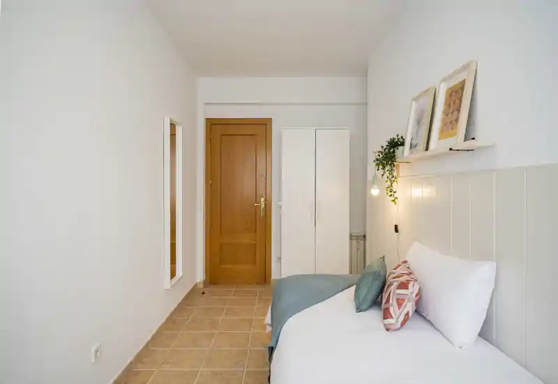 Camera da letto luminosa e stretta con un letto singolo ben rifatto, cuscini decorativi, mensola con quadri e una lampada a sospensione; armadio semplice e specchio a figura intera vicino alla porta in legno.