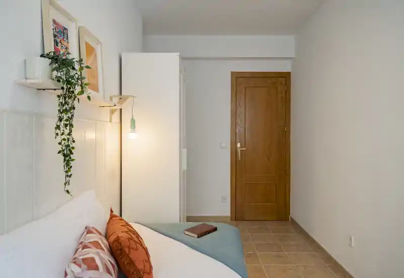 Dormitorio luminoso y acogedor con cama arreglada con cojines decorativos y una manta azul verdosa, lámpara colgante cálida y armario blanco junto a la puerta de madera.