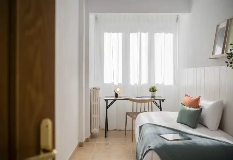 Bright, modern single bedroom with a neatly made bed, decorative cushions and a small desk by a sunlit window — ideal for a cozy, work-friendly stay.