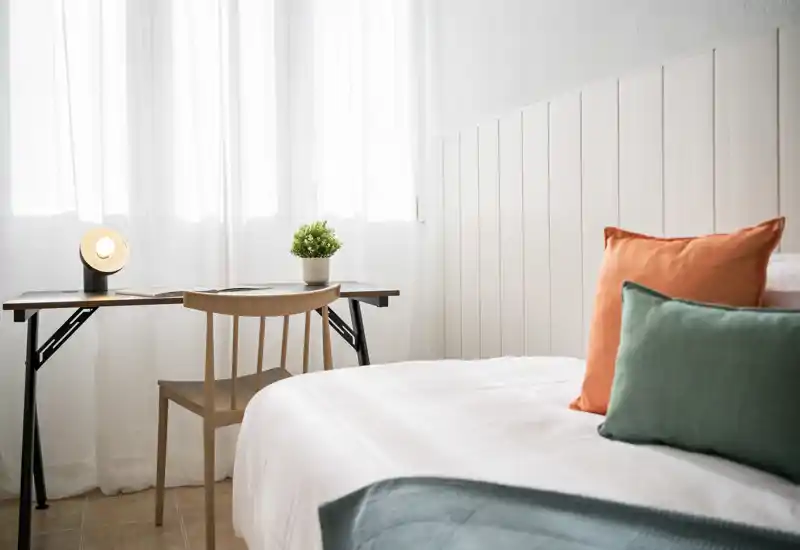 Bright, minimal bedroom with a neatly made bed accented by colorful cushions and a small desk by the window — ideal for relaxing or working.