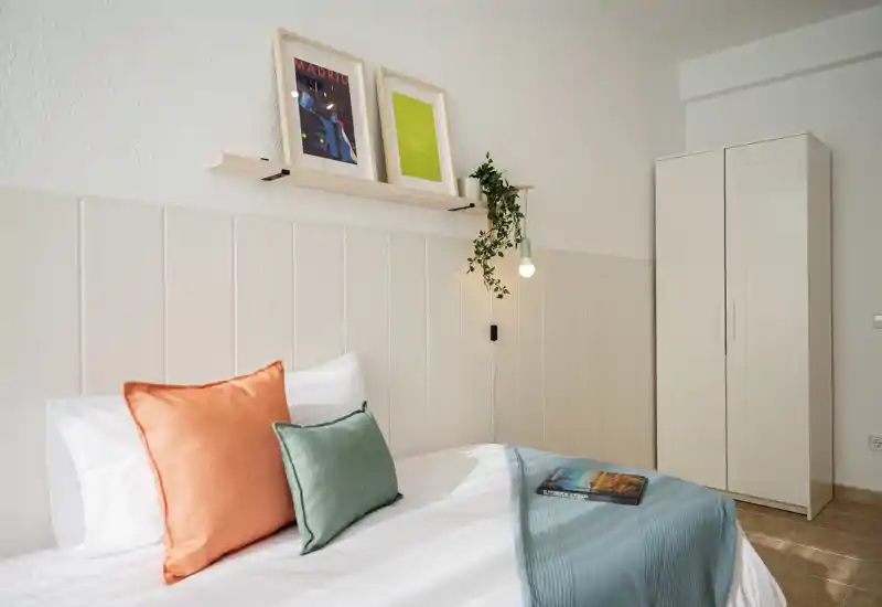 Bright, modern bedroom with a neatly made bed, colorful cushions and a wardrobe — styled with a hanging plant and framed art for a welcoming feel.