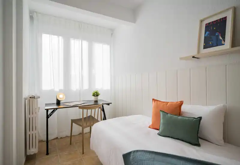 Bright, compact bedroom with a comfortable single bed dressed in white linen, colourful accent cushions and a small desk by the window — ideal for working with natural light.