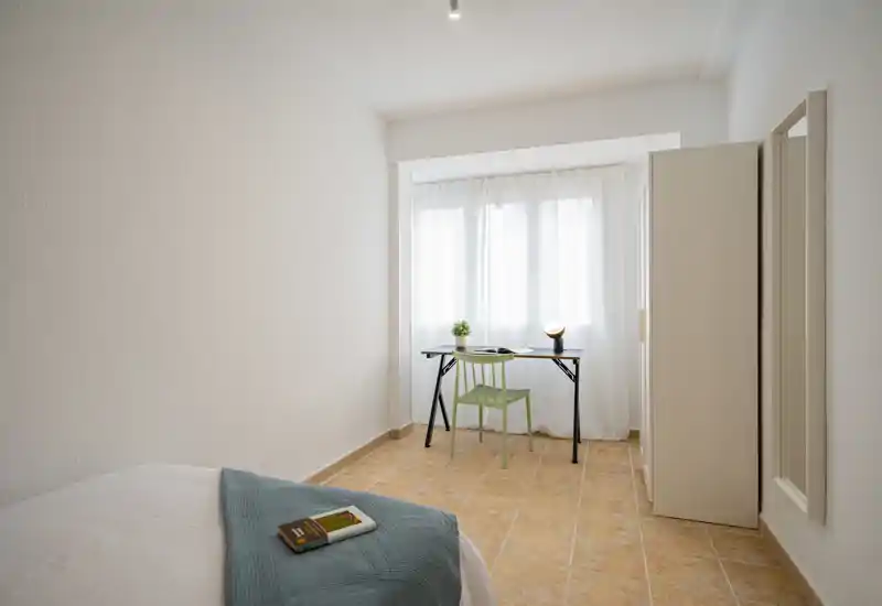Bright minimal bedroom with a neatly made bed in the foreground, a small desk by a sunlit window and a wardrobe—calm, airy and ready for guests.