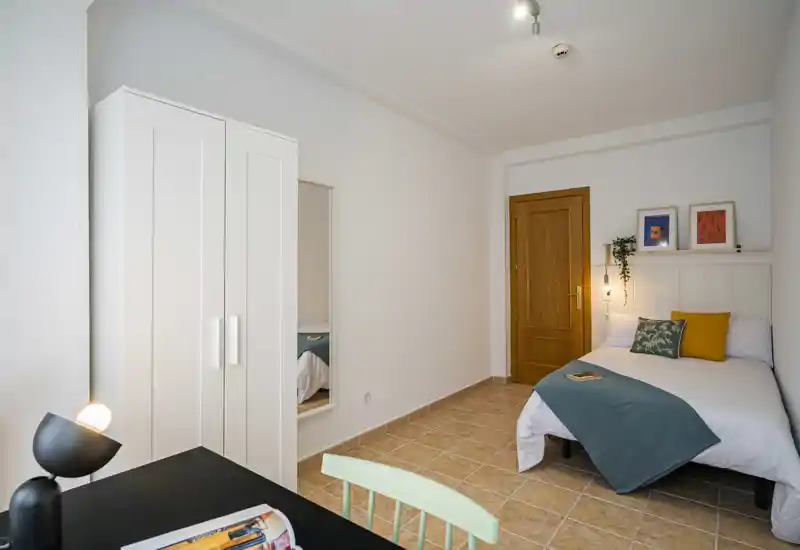 Bright, neatly staged bedroom with a double bed dressed in white linens and a teal throw, bedside shelf with framed prints and a hanging lamp, wardrobe and desk visible — ideal for rental photos.