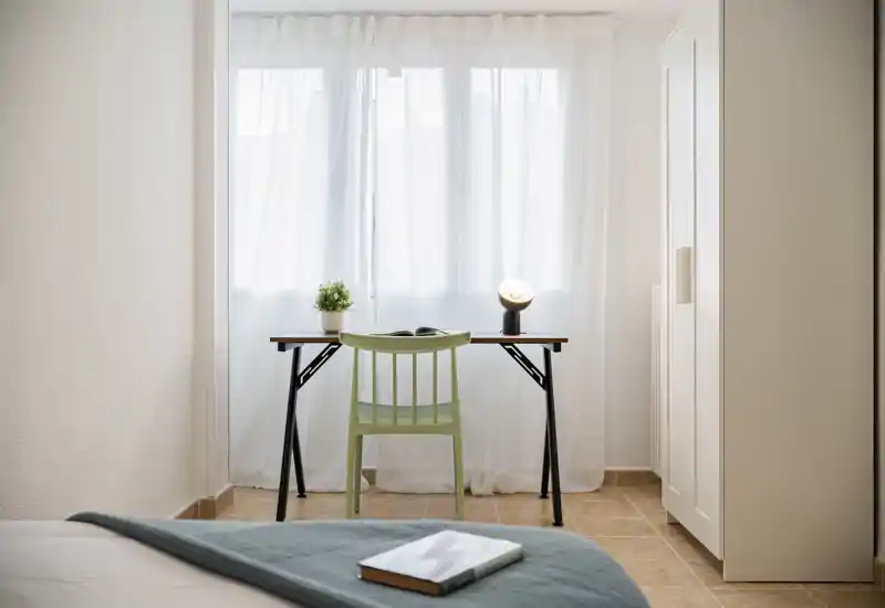 Bright, minimal bedroom corner with a bed in the foreground, a simple desk and chair by a sheer-curtained window, plus a wardrobe to the right — ideal for a restful stay or remote work.