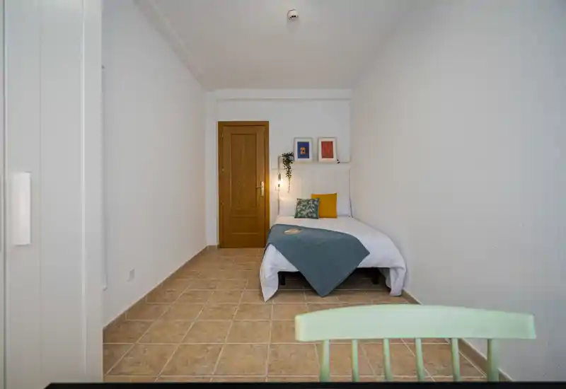 Compact, tidy bedroom with a single bed dressed in white linen, colorful cushions and a teal throw; warm wall lamp and simple artwork create a cozy atmosphere.