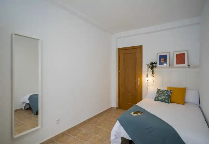 Dormitorio sencillo y ordenado con una cama individual vestida en ropa blanca y una manta verde azulado, cojines decorativos, cuadros apoyados y un espejo en la pared que refleja parte de la cama. Paredes neutras y suelo de baldosas cálidas crean un ambiente tranquilo y minimalista.