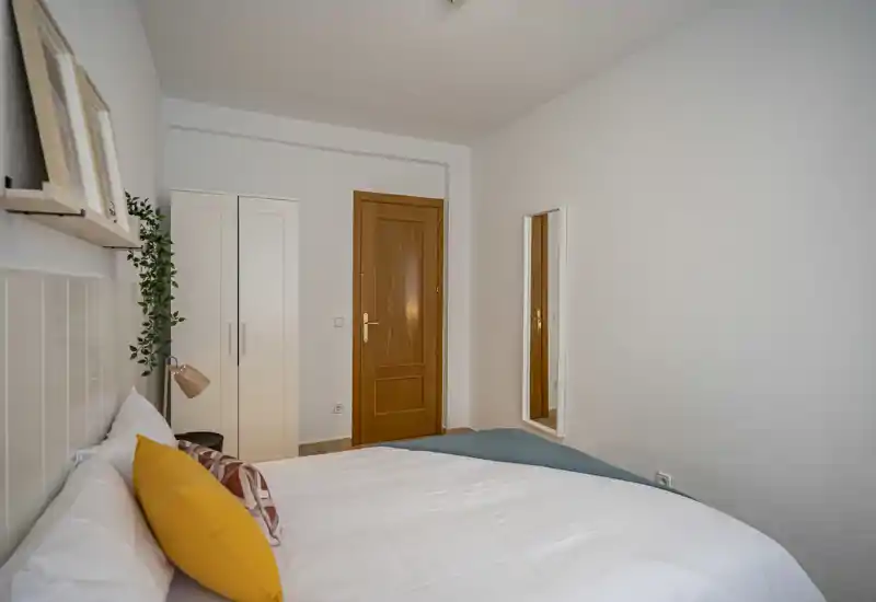 Well-lit, simply styled bedroom with a large bed dressed in white linens, decorative pillows and a small wardrobe near the door; neutral walls and warm wooden door add a cozy touch.