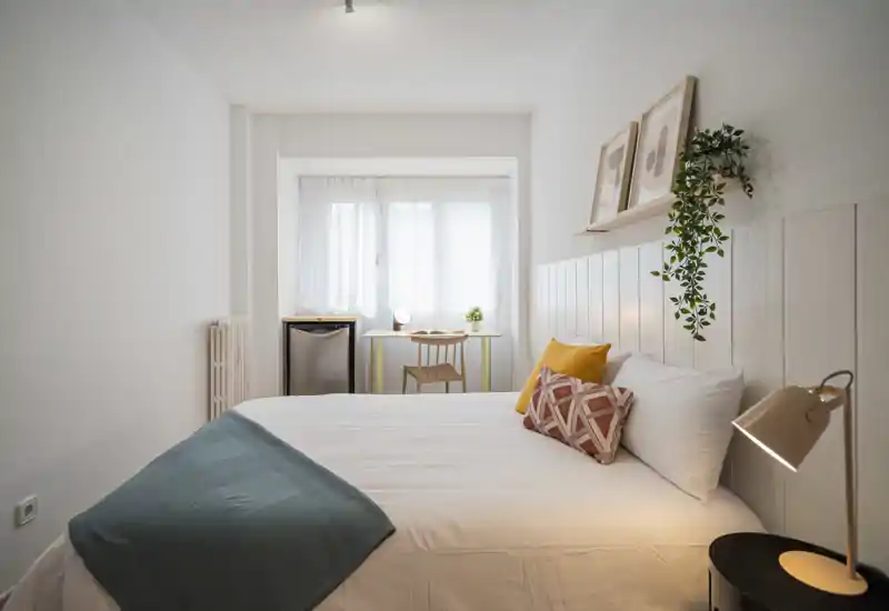 Bright, modern bedroom with a neatly made double bed, decorative cushions, a bedside lamp and a small desk by the window — ideal for restful stays.