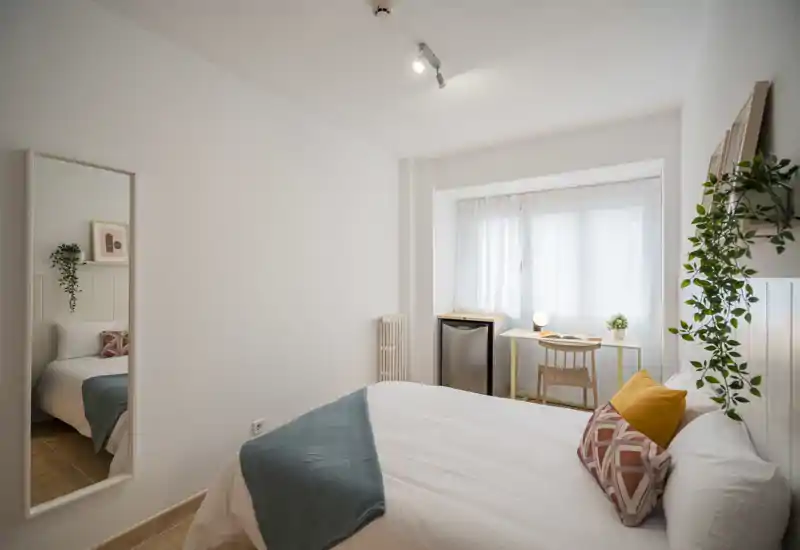 Bright, well-styled bedroom with a comfortable double bed, decorative cushions and a small desk by the window. Natural light and clean minimalist decor create a calm, welcoming atmosphere.