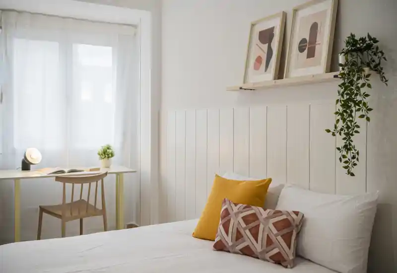 Camera da letto luminosa e accogliente con letto ben rifatto, cuscini decorativi, pianta sospesa e una piccola scrivania vicino alla finestra con tenda voile.