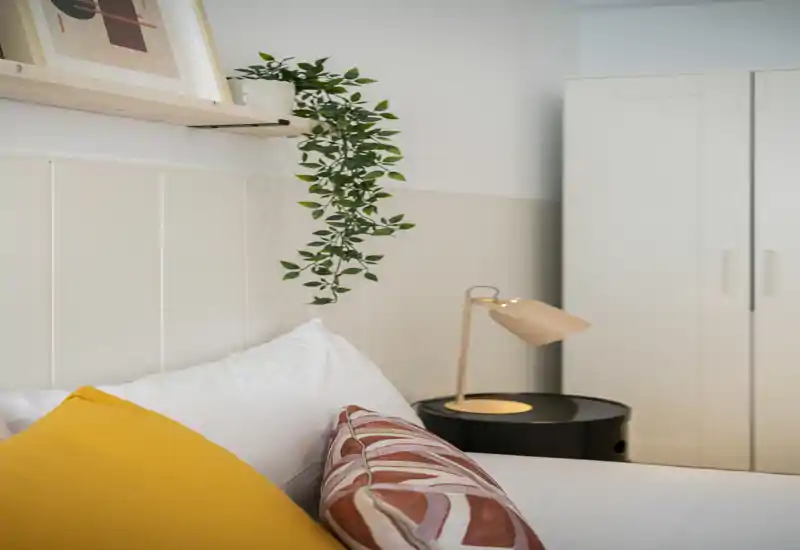 Bright, styled bedroom corner featuring a neatly made bed with decorative pillows, a bedside lamp and a wardrobe in the background — cozy, modern and well-lit.