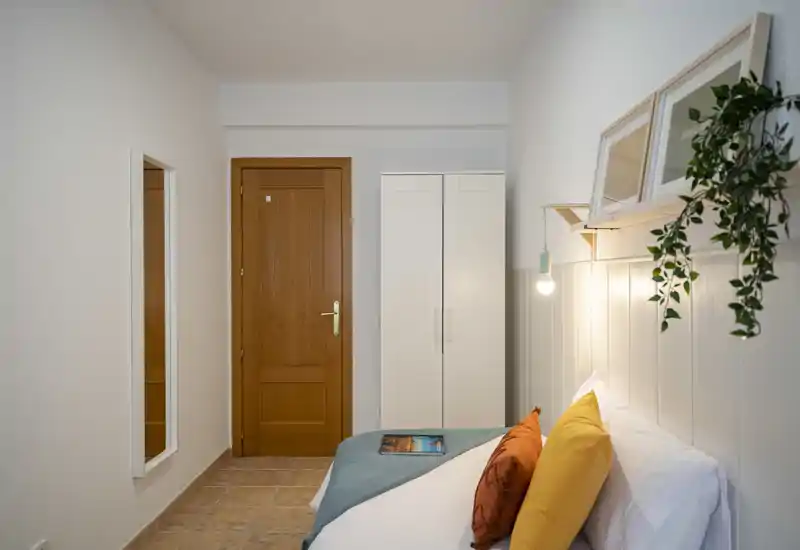 Dormitorio luminoso y bien dispuesto con cama cómoda, cojines de colores, lámpara colgante junto a la cama y un armario sencillo — perfecto para una estancia acogedora.