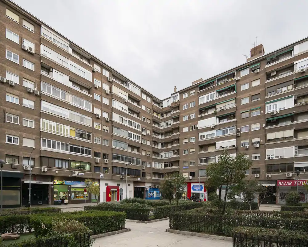 Urban courtyard with mid-rise residential blocks, ground-floor shops and planted hedges — a calm communal outdoor space.