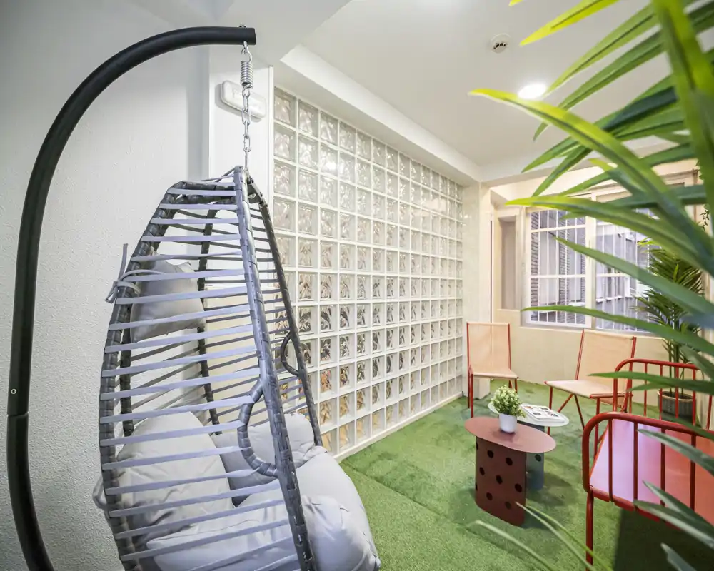 Covered balcony/indoor terrace with green artificial turf, hanging rattan swing chair and colorful seating — a bright, casual spot for relaxing or reading.