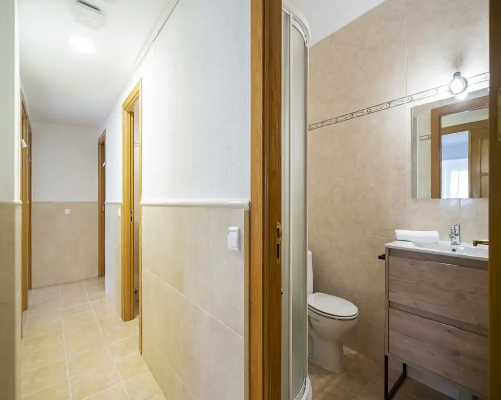 Bright tiled corridor with several doorways and an adjacent small bathroom featuring a shower, vanity and toilet. Clean, neutral finishes suitable for a rental listing.