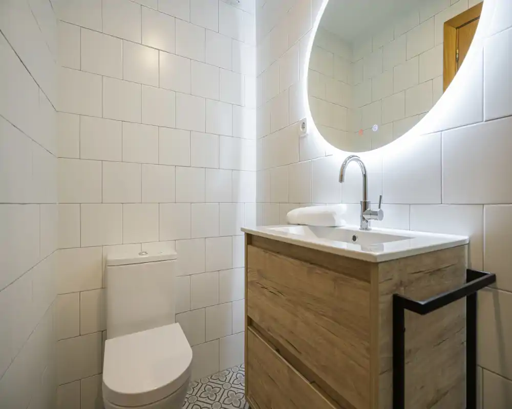 Modern small bathroom with a wall-mounted illuminated round mirror, white tiled walls, a wooden vanity with sink and a toilet; bright, clean and minimal.