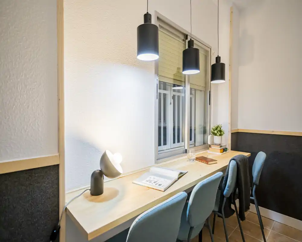 Bright, modern study/workspace with a long wooden desk, three chairs, pendant lights and a desk lamp — ideal for focused work or study.