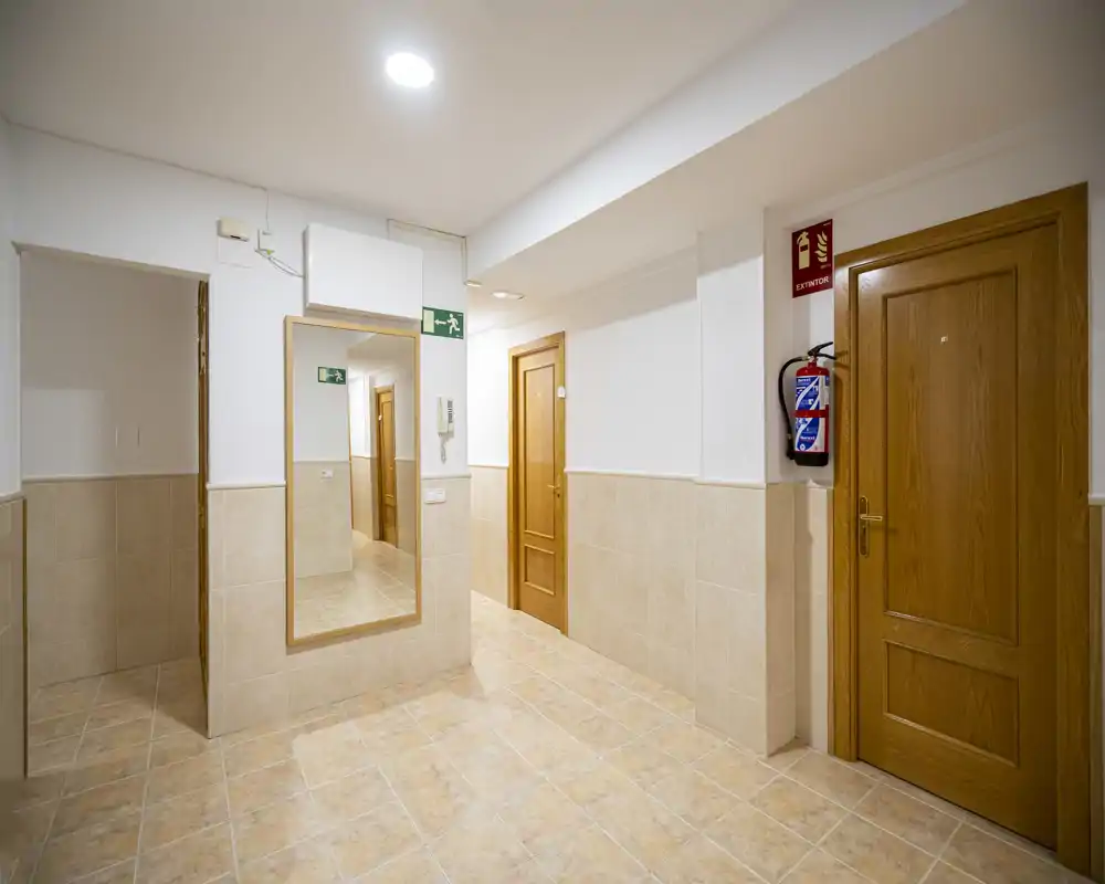 Well-lit apartment corridor with tiled floor, wooden doors and safety signage. Neutral, clean space suitable for entrance or common hallway.