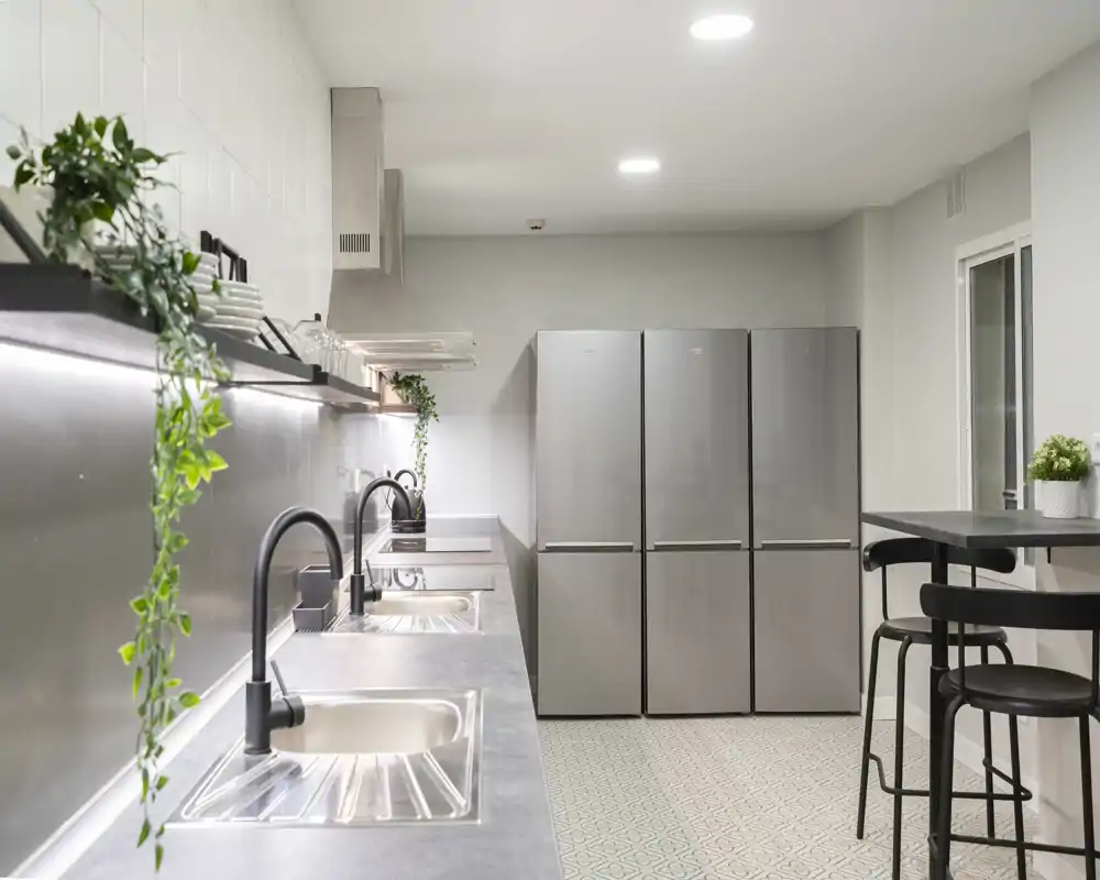 Modern shared kitchen with a long stainless counter, multiple sinks and faucets, three tall stainless fridges and a small high table with stools. Clean, well-lit and styled with simple greenery for a contemporary communal cooking area.