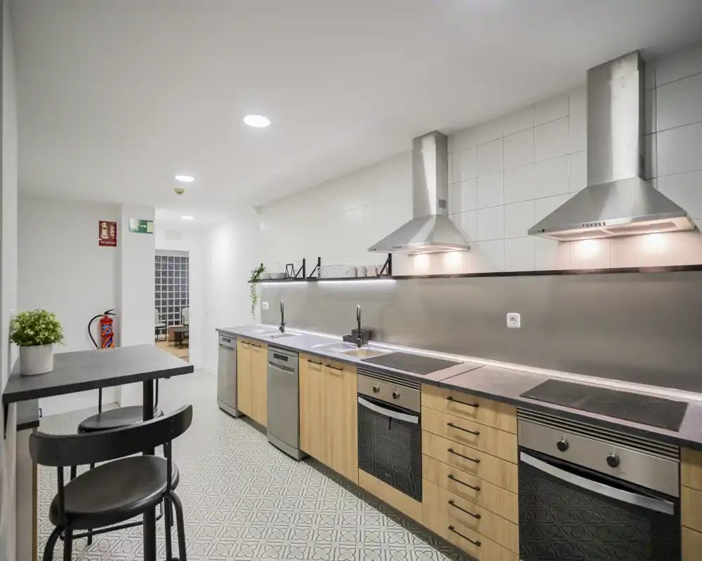 Modern fully equipped kitchen with long countertop, built-in ovens, induction hobs and stainless steel range hoods; light wood cabinets and patterned floor tiles create a clean, contemporary look.