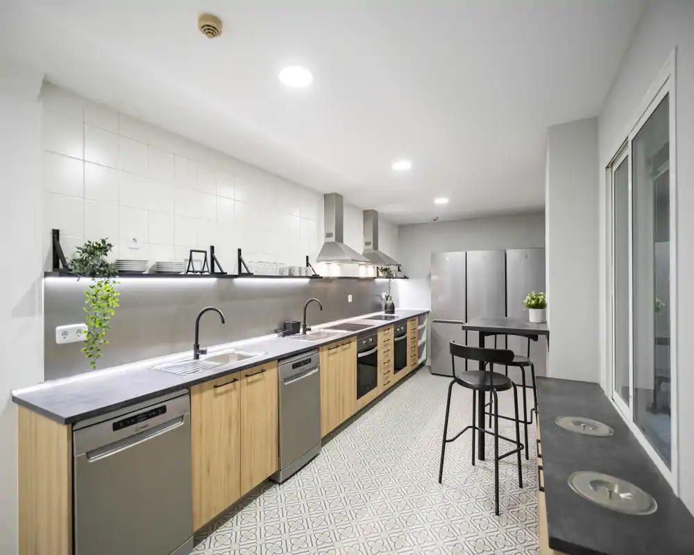 Bright modern kitchen with a long wooden cabinet run, stainless-steel appliances and a tiled floor — ideal for shared or commercial-style cooking spaces.