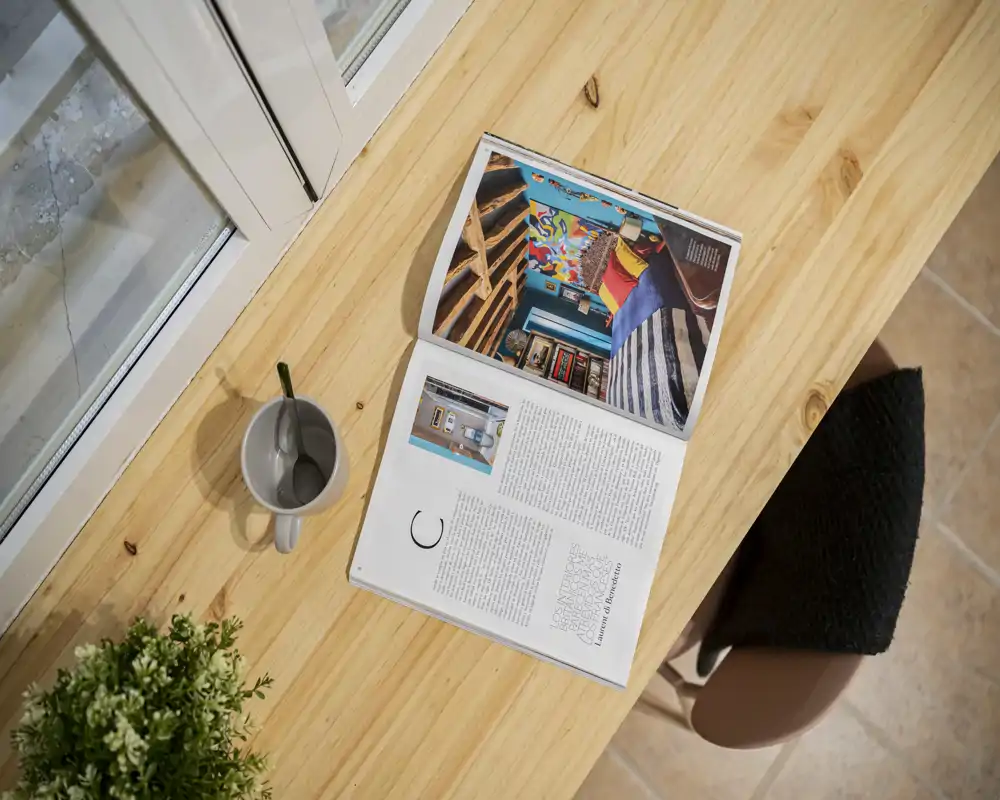 Neat wooden table by a window with a magazine, mug and a plant—ideal close-up shot for a dining or breakfast nook showcasing natural light and a cozy casual setup.