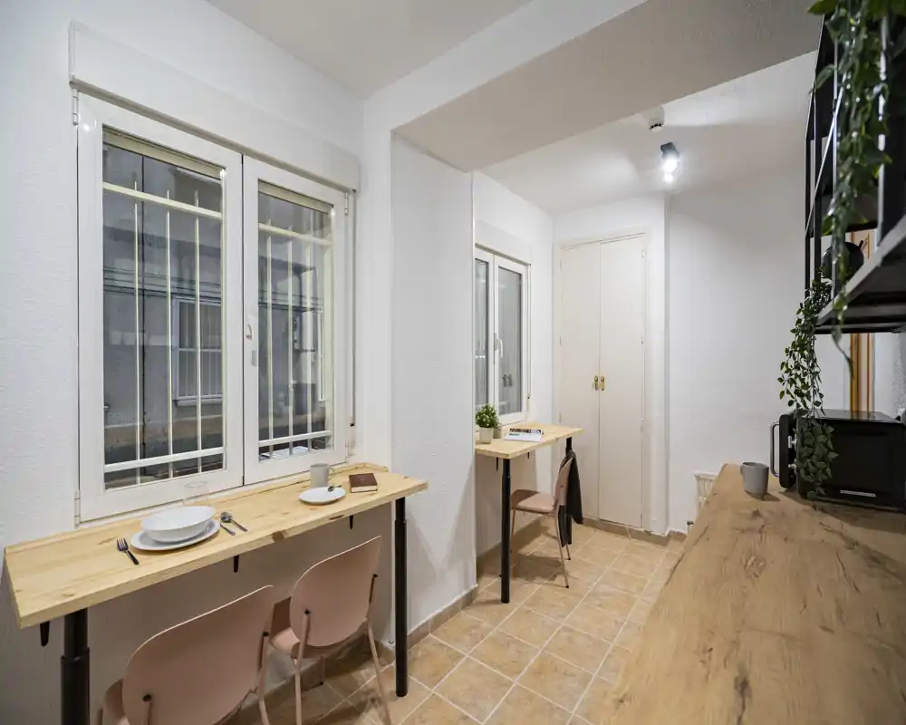Compact, narrow living/dining area with two breakfast bars by the windows, simple pink chairs and wooden surfaces. Bright, functional layout ideal for a small urban rental.