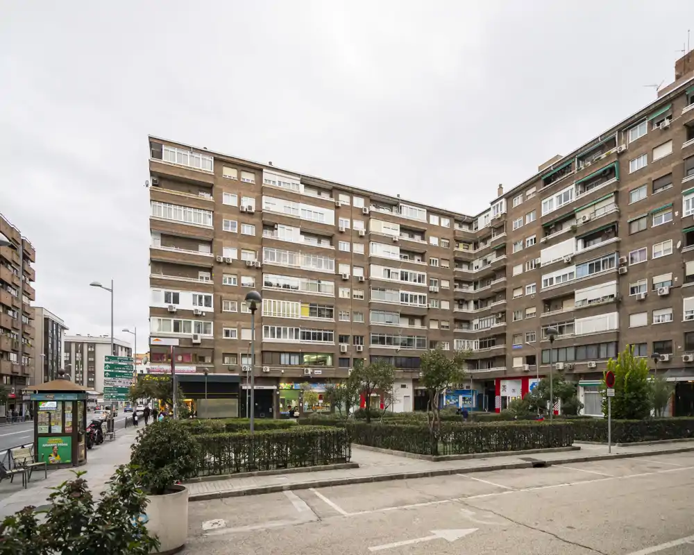 Street-facing view of a multi-storey residential building with small planted squares and ground-floor shops, suitable for showcasing the building exterior and neighborhood.