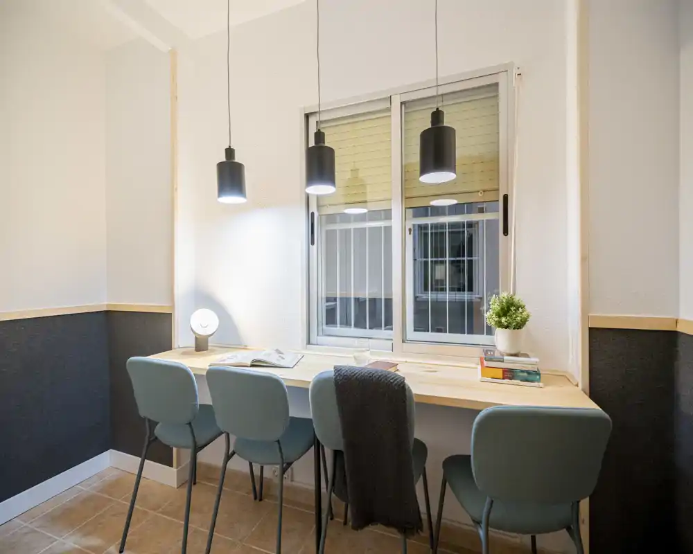 Bright, modern workspace with a long wooden desk beneath a window, four stylish chairs, pendant lights and small decorative plant — ideal for shared study or remote work.