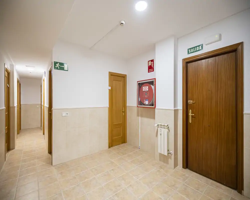 Well-lit apartment corridor/entrance hall with tiled floor, wooden doors and visible emergency signage — clean and functional common area.