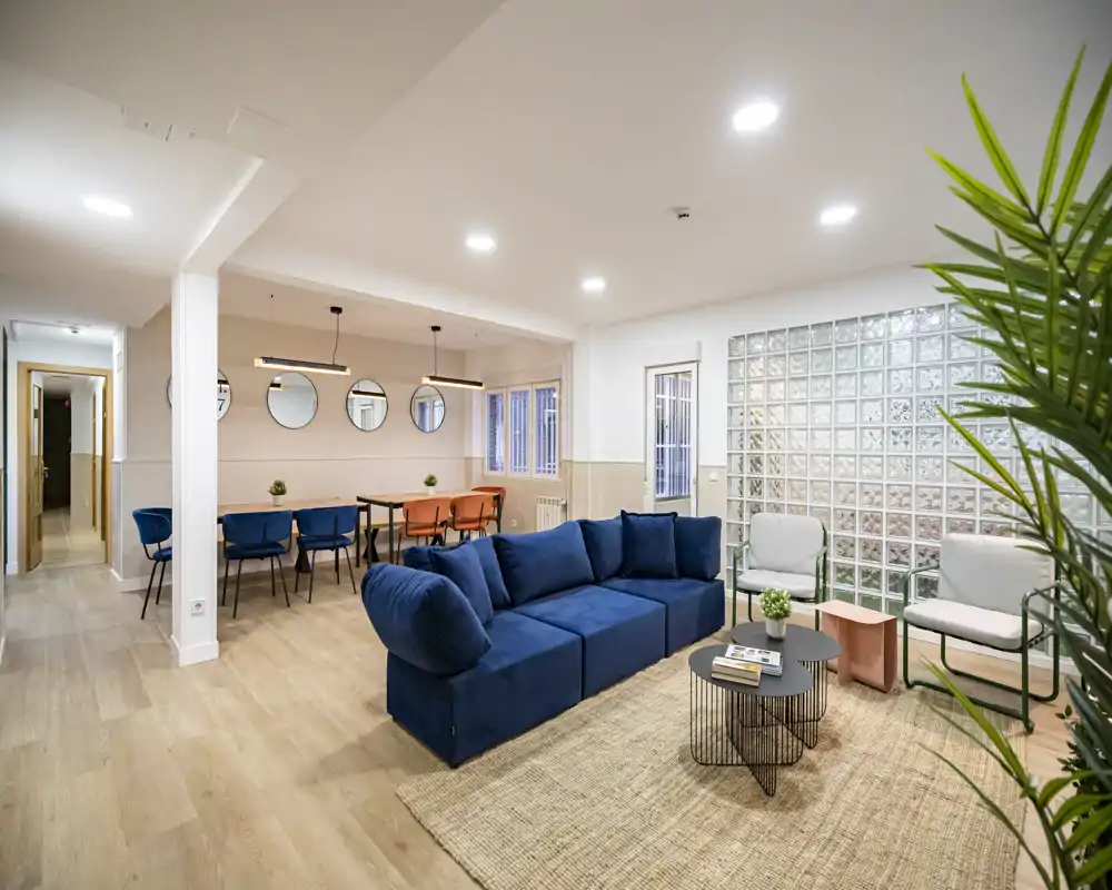 Spacious, modern living room with a deep blue modular sofa, woven rug, two armchairs and a coffee table; bright, well-staged open space with dining area visible in the background.