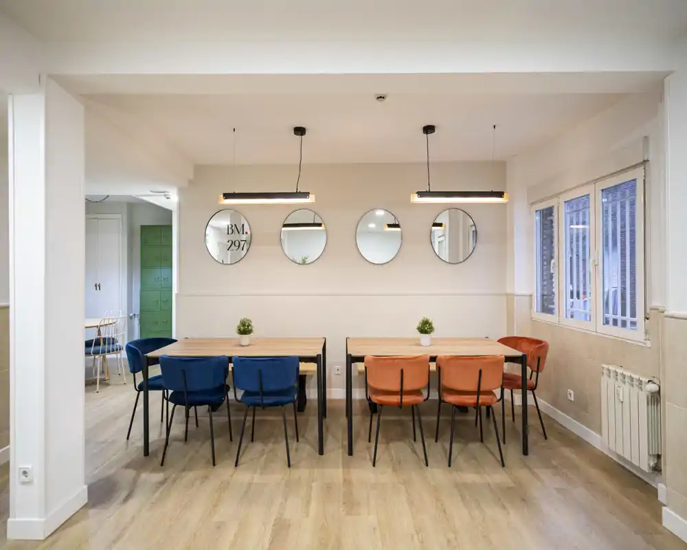 Modern dining area with two wooden tables and colorful upholstered chairs (blue and orange), pendant lights and decorative round mirrors — bright, inviting space suitable for shared meals or co-working.