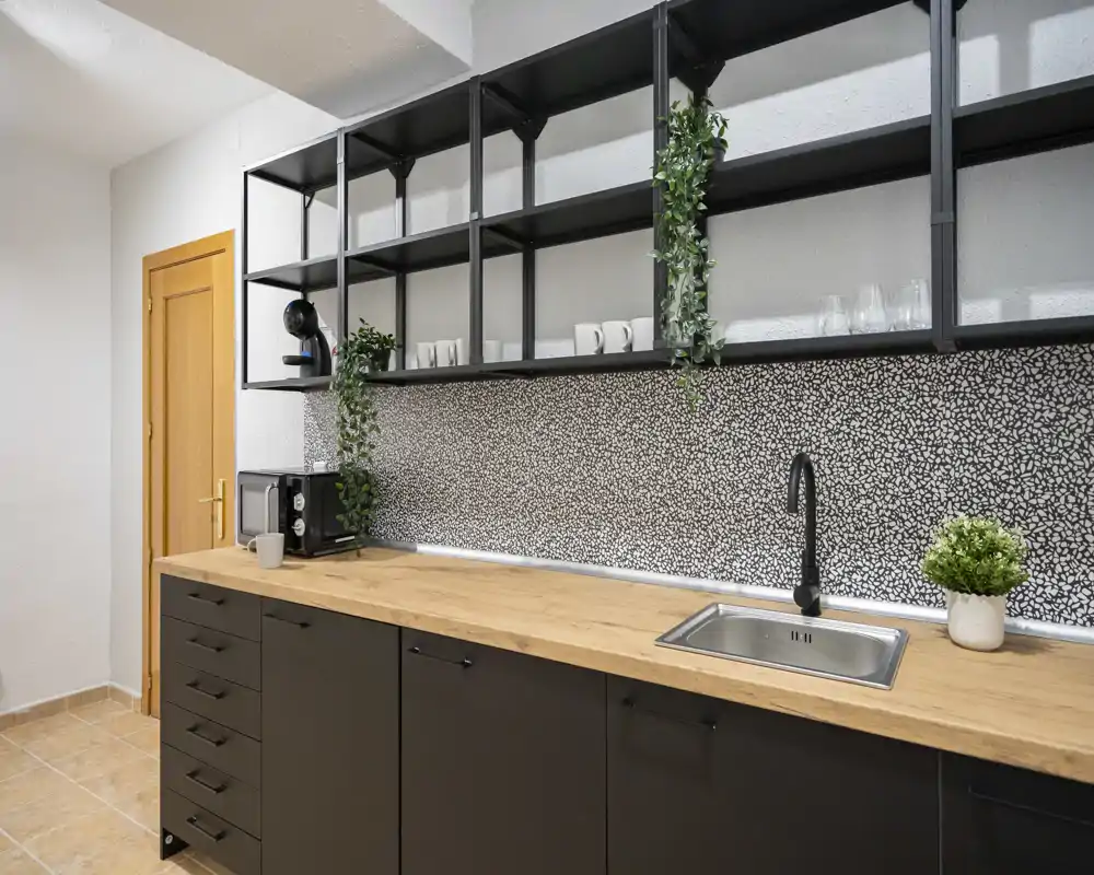 Modern compact kitchen with a wooden countertop, black lower cabinets, open shelving and a patterned backsplash; sink and small appliances visible, bright and neatly styled.