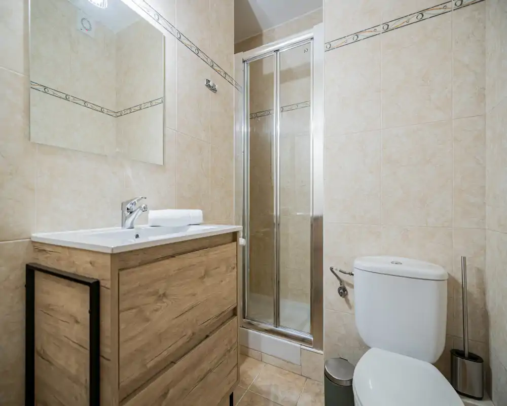 Clean, compact bathroom with a wooden vanity, sink and a glass-enclosed shower. Neutral beige tiles and a wall mirror create a bright, tidy feel.