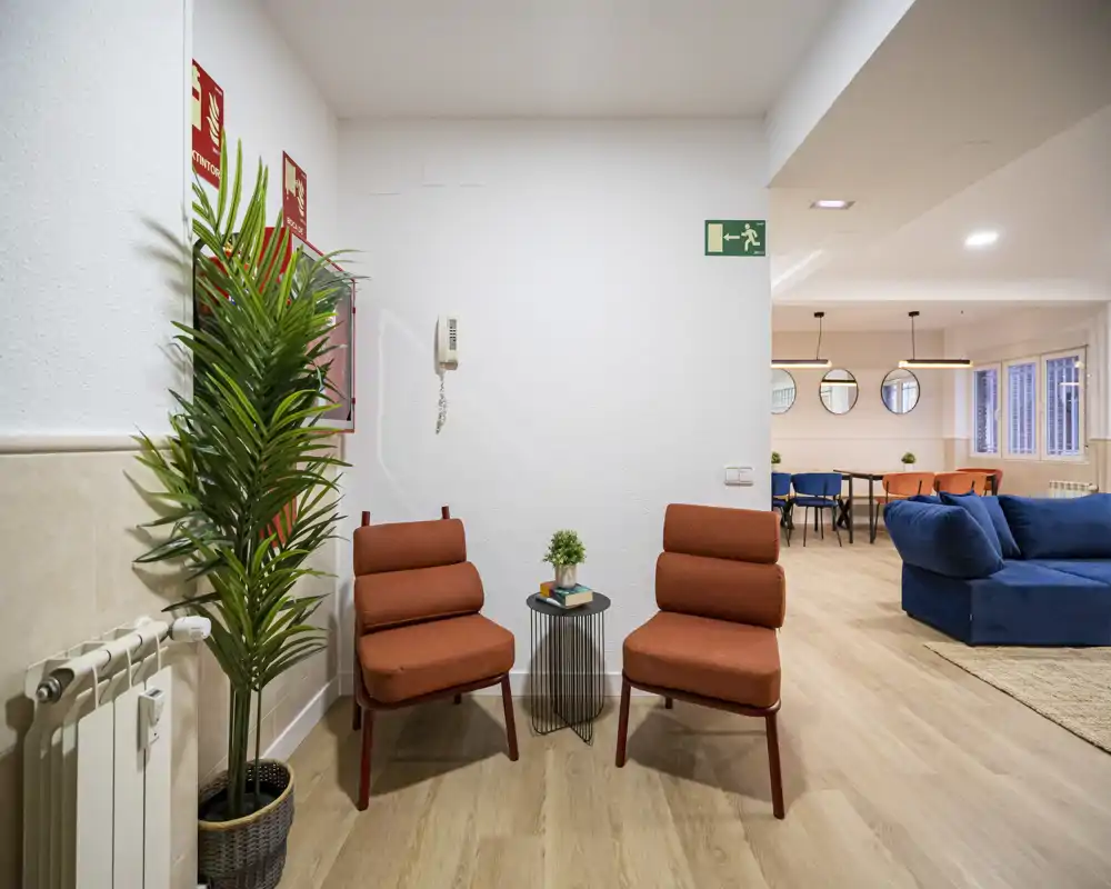 Bright, modern communal area with comfortable seating: two rust-colored lounge chairs, a blue sofa and a dining area visible in the background. Ideal for socializing or relaxing in a stylish shared living space.