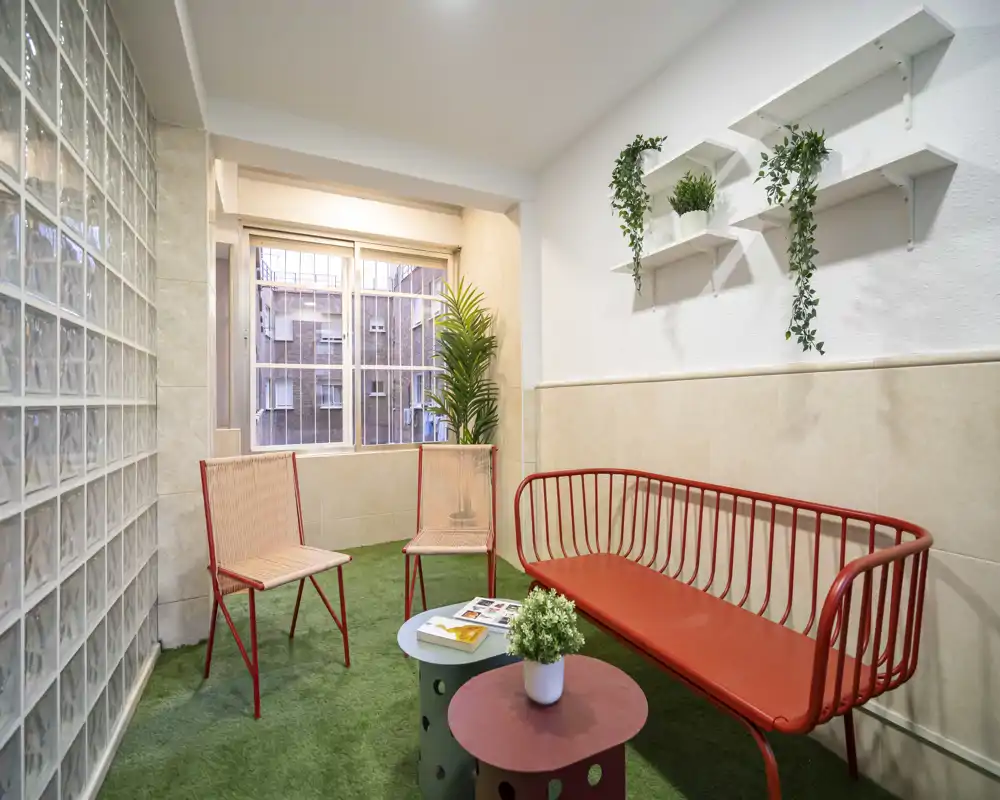 Small bright living/lounge area with red metal seating, a pair of chairs, low coffee tables and decorative plants, set on green turf-like flooring near a window.