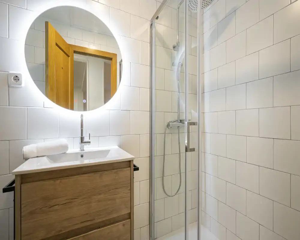 Bright, modern bathroom with a glass shower enclosure, illuminated round mirror and wooden vanity with sink — clean and contemporary styling ideal for rental listings.